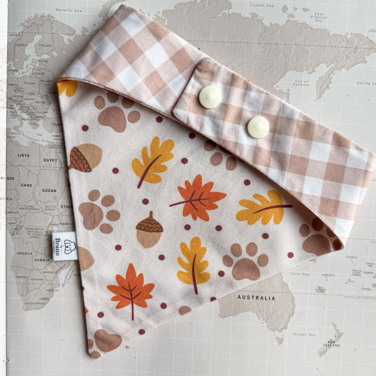 A dog bandana laid flat on a vintage world map. The bib has an off-white background with a pattern of autumn leaves, acorns, paw prints, and dots in orange, mustard yellow, brown, and muted red. On the photo there is the other side shown with light brown gingham print and two plastic pearl buttons.
