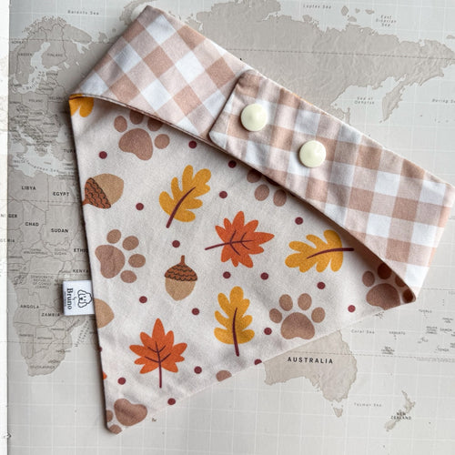 A dog bandana laid flat on a vintage world map. The bib has an off-white background with a pattern of autumn leaves, acorns, paw prints, and dots in orange, mustard yellow, brown, and muted red. On the photo there is the other side shown with light brown gingham print and two plastic pearl buttons.