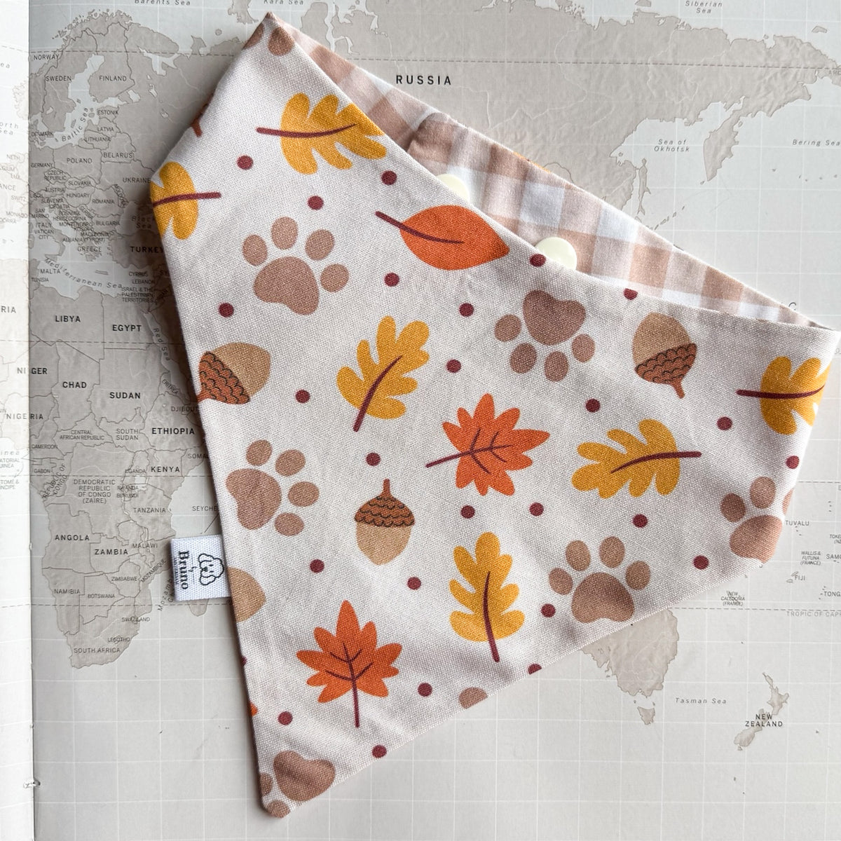 A dog bandana laid flat on a vintage world map. The bandana has an off-white background with a pattern of autumn leaves, acorns, paw prints, and dots in orange, mustard yellow, brown, and muted red.