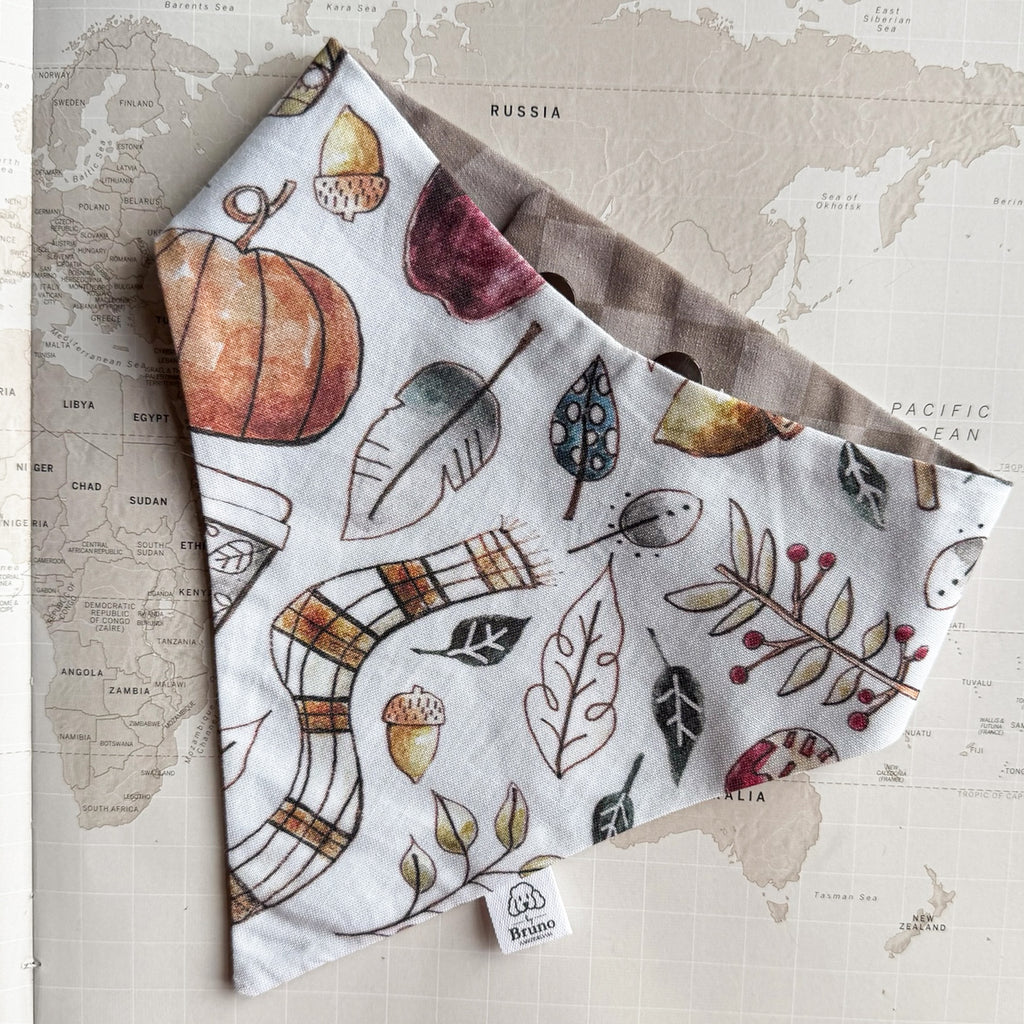 A dog bandana with an autumn watercolor print, laid flat on a vintage-style world map background. The print features pumpkins, acorns, leaves, and a plaid scarf pattern on a white base.