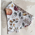A dog bandana with an autumn watercolor print, laid flat on a vintage-style world map background. The print features pumpkins, acorns, leaves, and a plaid scarf pattern on a white base.