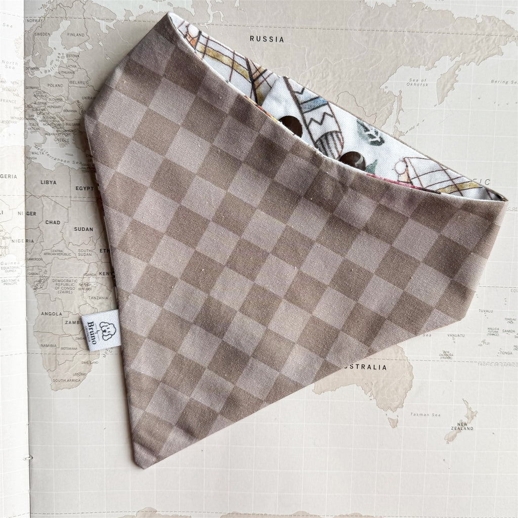 A dog bandana with a taupe and beige checkerboard pattern on the outside, and a white patterned lining with small illustrated figures, laid flat on a vintage-style world map background.