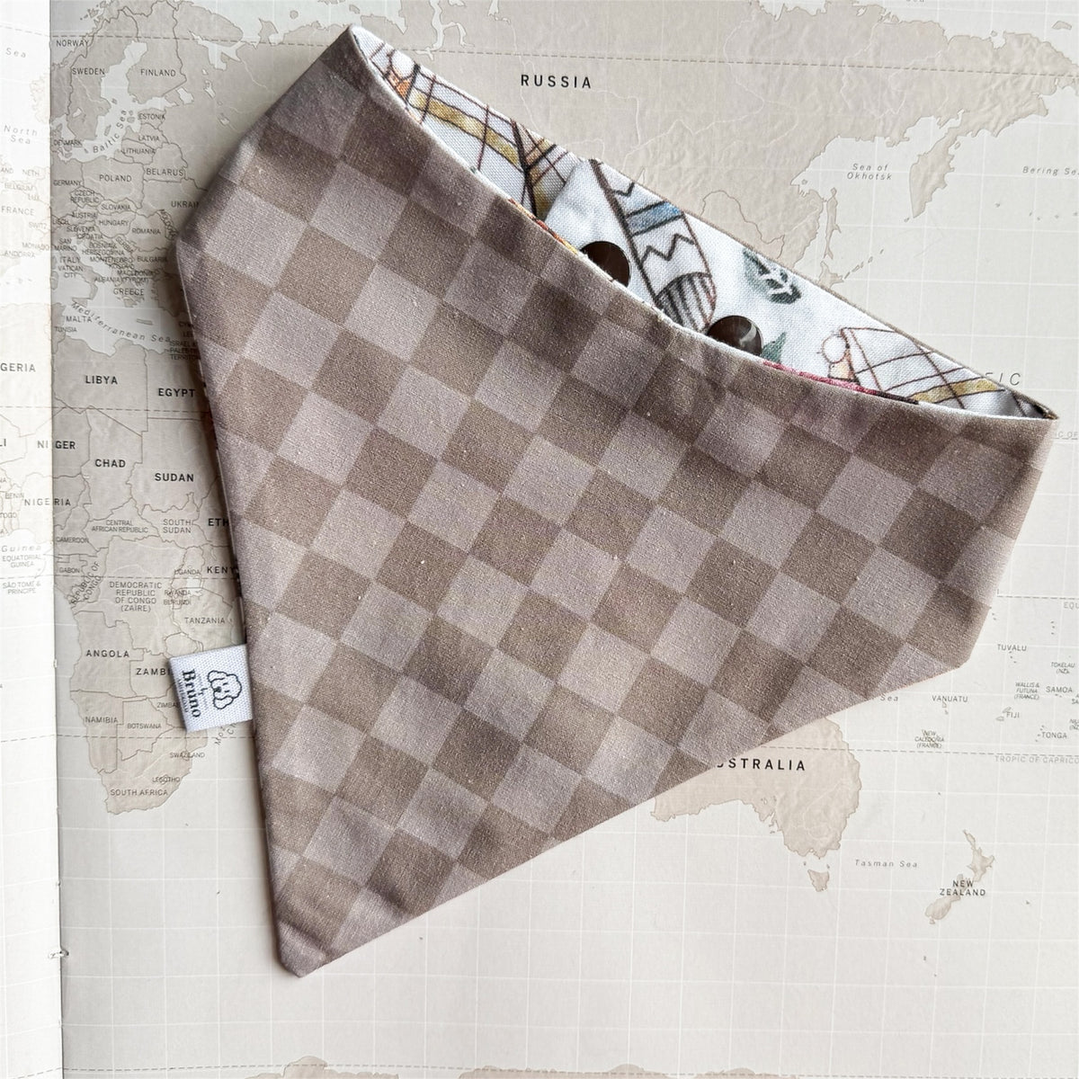 A dog bandana with a taupe and beige checkerboard pattern on the outside, and a white patterned lining with small illustrated figures, laid flat on a vintage-style world map background.
