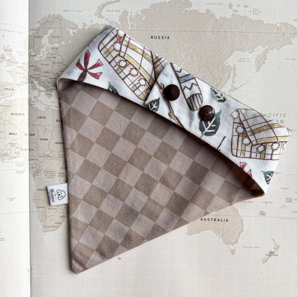 A dog bandana laid flat on a vintage world map background. The fabric is primarily taupe and beige checkered on one side, and the reverse side shows a white background with illustrated vintage buses and leaves.
