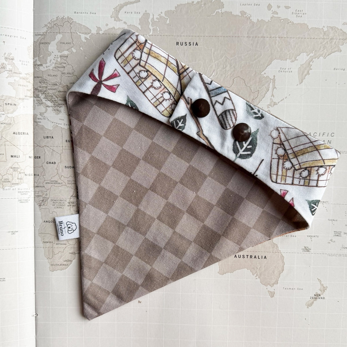 A dog bandana laid flat on a vintage world map background. The fabric is primarily taupe and beige checkered on one side, and the reverse side shows a white background with illustrated vintage buses and leaves.