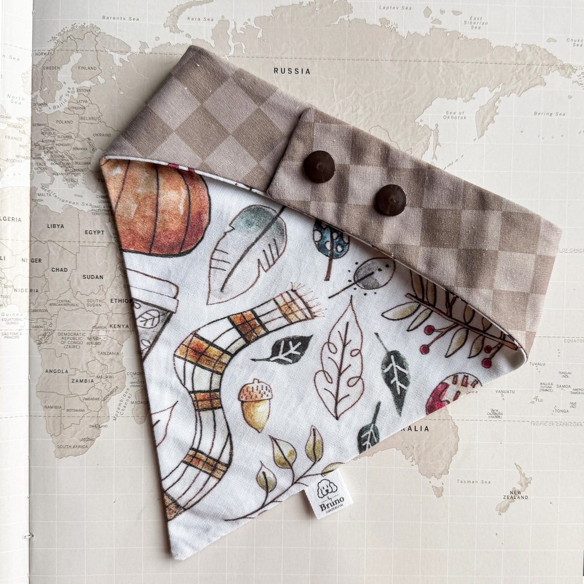 A dog bandana laid flat on a vintage world map background. The bandana has a taupe and cream checkerboard fold-over section with two dark brown buttons, and the main panel features an autumn-themed print with a snake, plaid scarf, leaves, acorns, and berries on a white base.