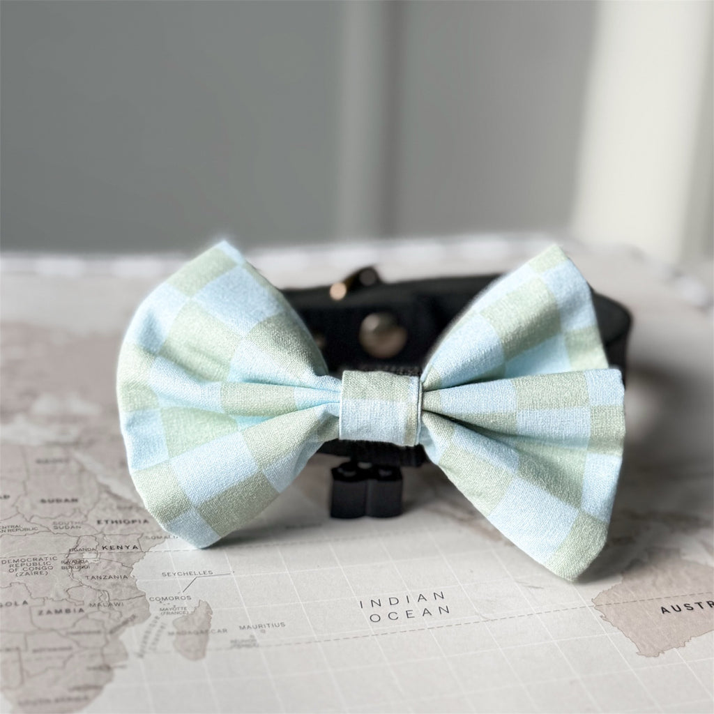 A light blue and mint green gingham bow tie displayed on a black stand, resting on a vintage-style world map.