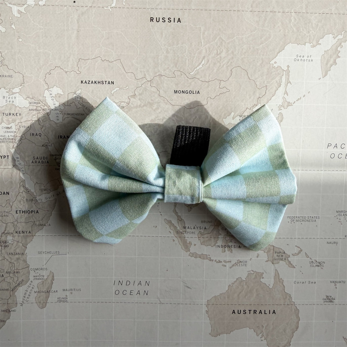 A light blue and mint green plaid bow tie laid flat on a vintage-style world map background.