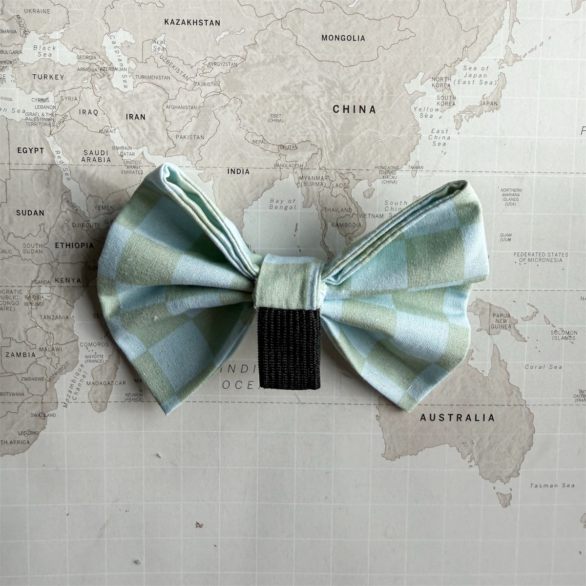 A dog bow tie with a light green and pale blue checkered pattern and a black center band, laid flat on a vintage-style world map background.