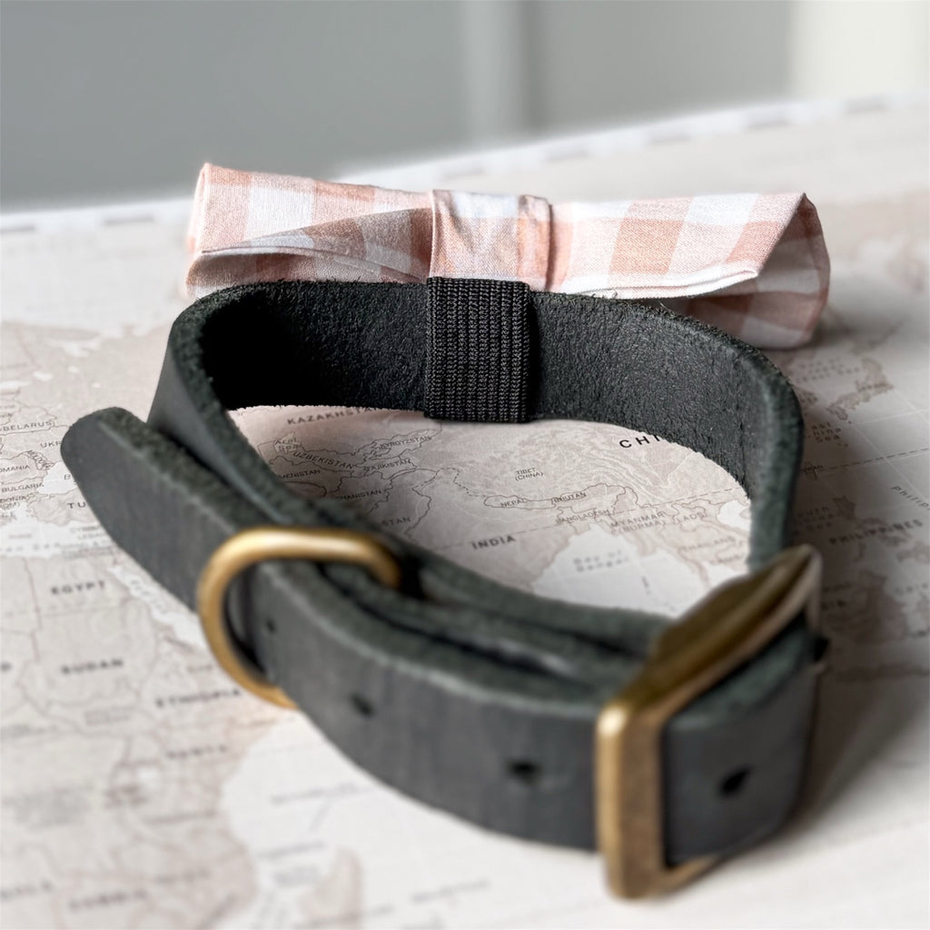 A dark charcoal gray leather belt with a brass buckle, styled on top of a vintage-style world map with a beige and white gingham bow tie behind it.