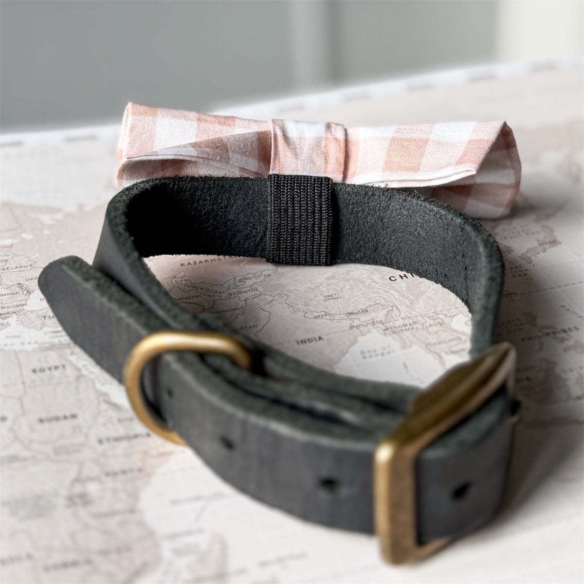A dark charcoal gray leather belt with a brass buckle, styled on top of a vintage-style world map with a beige and white gingham bow tie behind it.