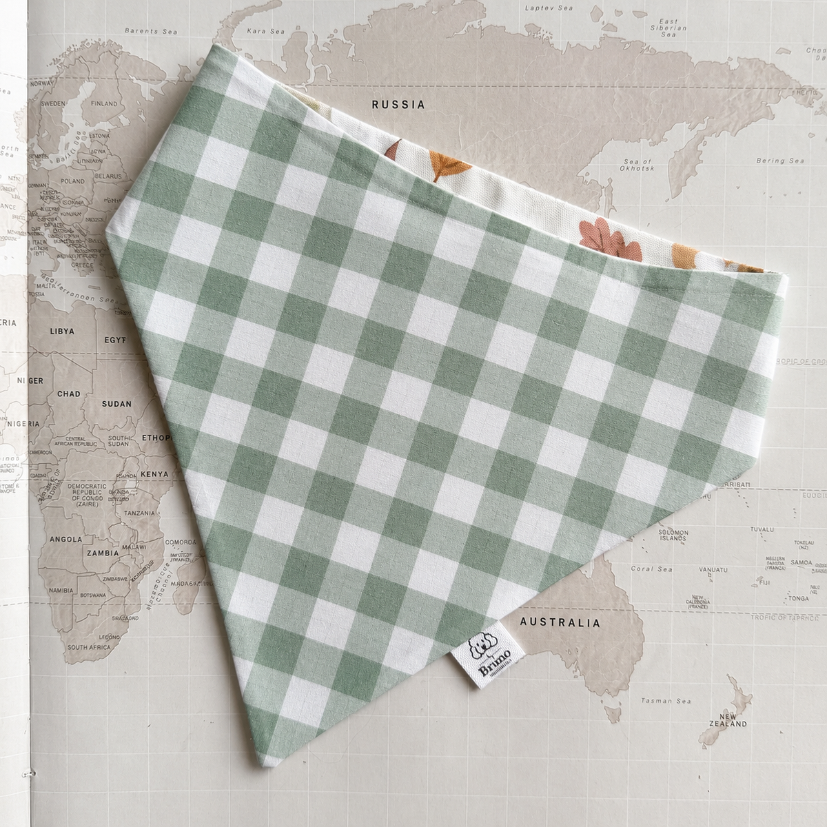 A green and white gingham check bandana with a small white sewn label, laid flat on a vintage-style world map background.