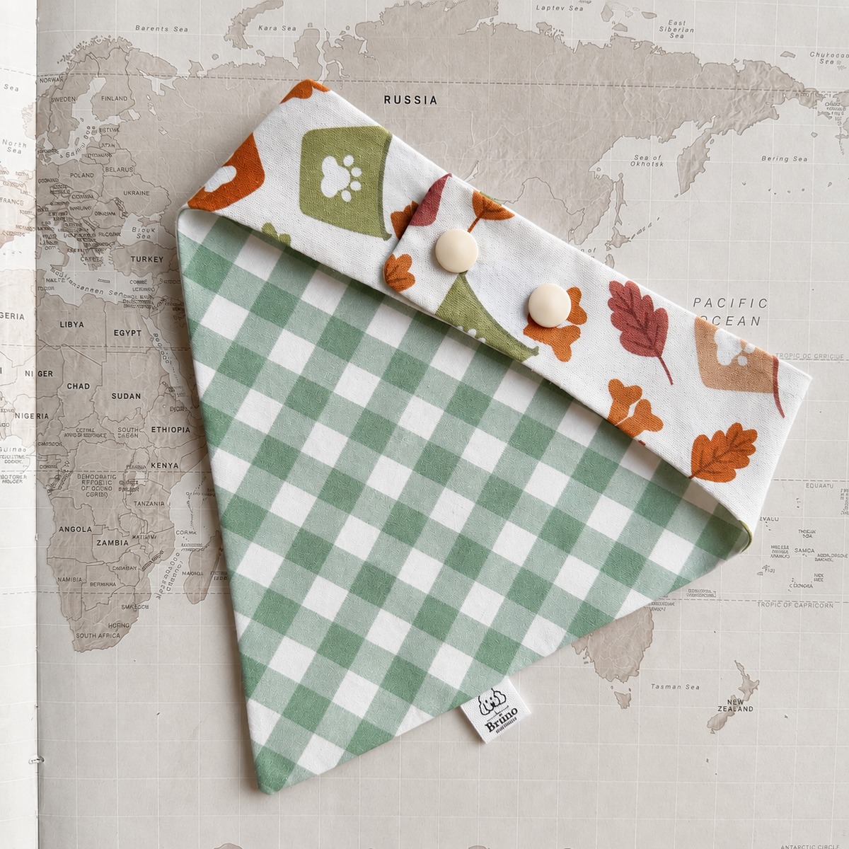 A green and white checkered triangular bandana with a folded flap showing an autumn leaf and paw print pattern, laid flat on a vintage world map background.