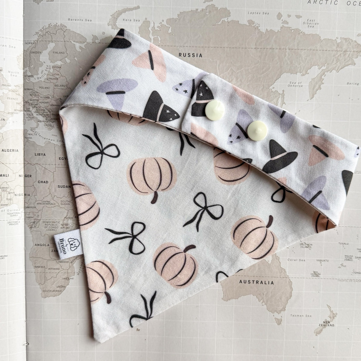 White cotton dog bandana with pastel orange pumpkins and black ribbons on a world map background. The photo is showing the back side with multi colored witch hats and two pearl plastic buttons.