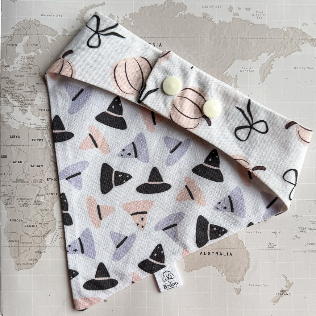 White cotton dog bandana with purple, orange and black witch hats on a world map background. The photo is showing the back side with pastel orange pumpkins and black ribbons  and two pearl plastic buttons.