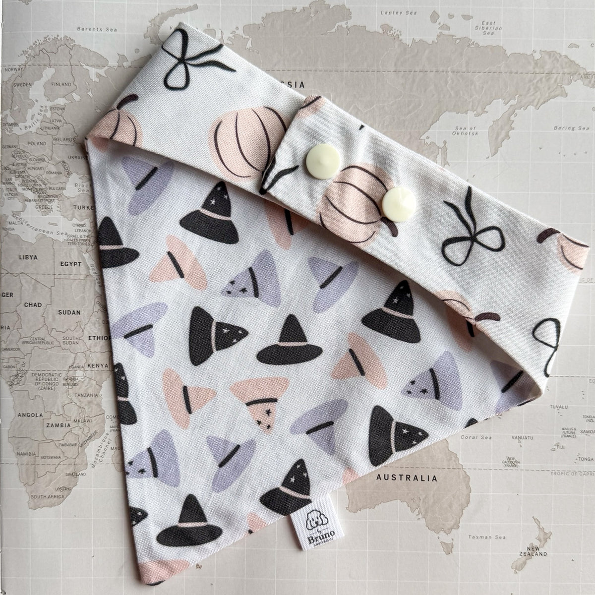 White cotton dog bandana with purple, orange and black witch hats on a world map background. The photo is showing the back side with pastel orange pumpkins and black ribbons  and two pearl plastic buttons.