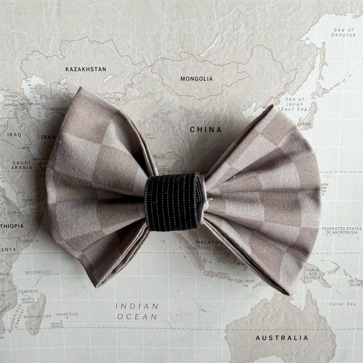 A checkered bow tie in a muted taupe-gray and beige pattern, featuring a black striped center band, displayed flat on a vintage-style world map background.