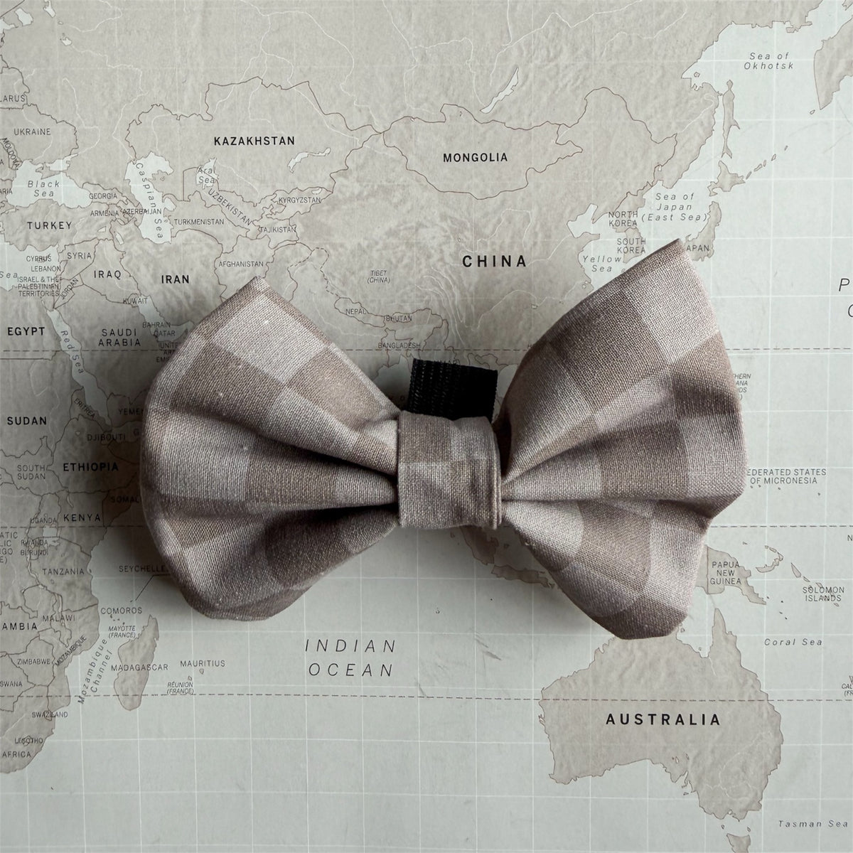 A dog bow tie with a muted taupe and beige checkered pattern, laid flat on a vintage-style world map background.