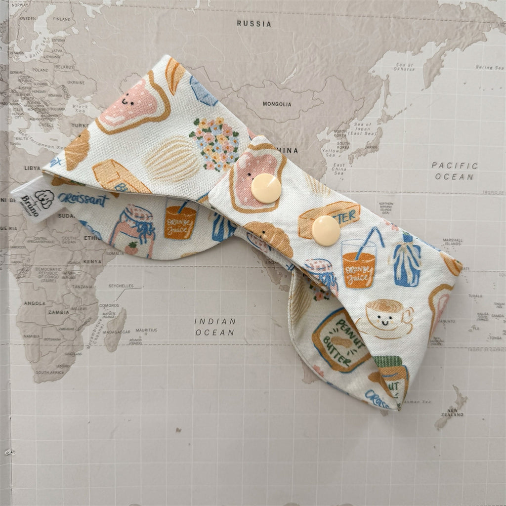 Back side of the dog bow bandana with plastic beige buttons and breakfast-themed motifs on a world map background.