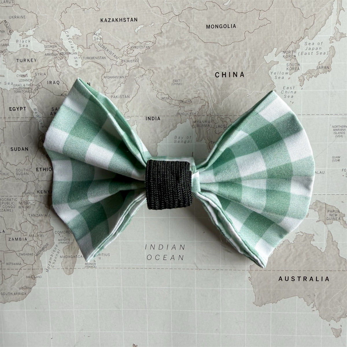 A green and white gingham check bow tie with a black elastic center band, laid flat on a vintage-style world map background.