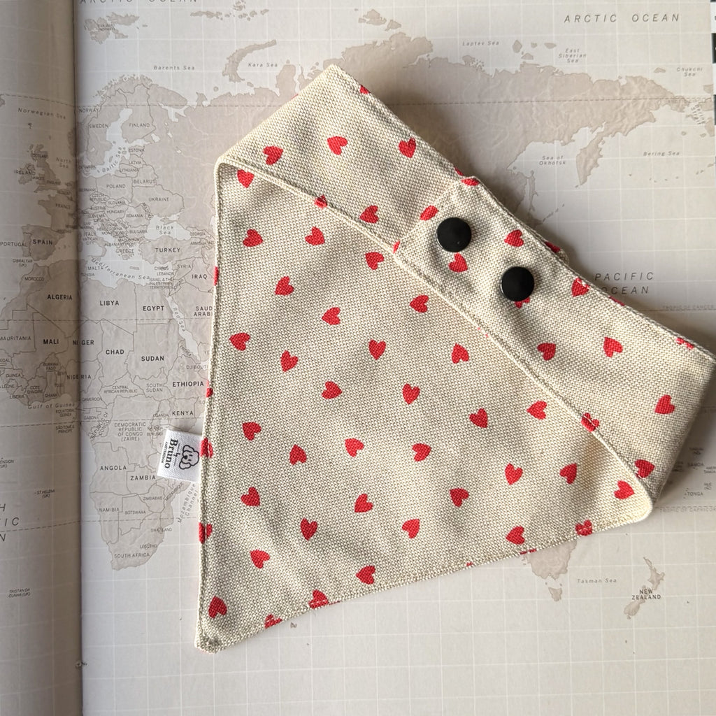 Back side of a beige dog bandana with red hearts and two black plastic buttons on a world map background.