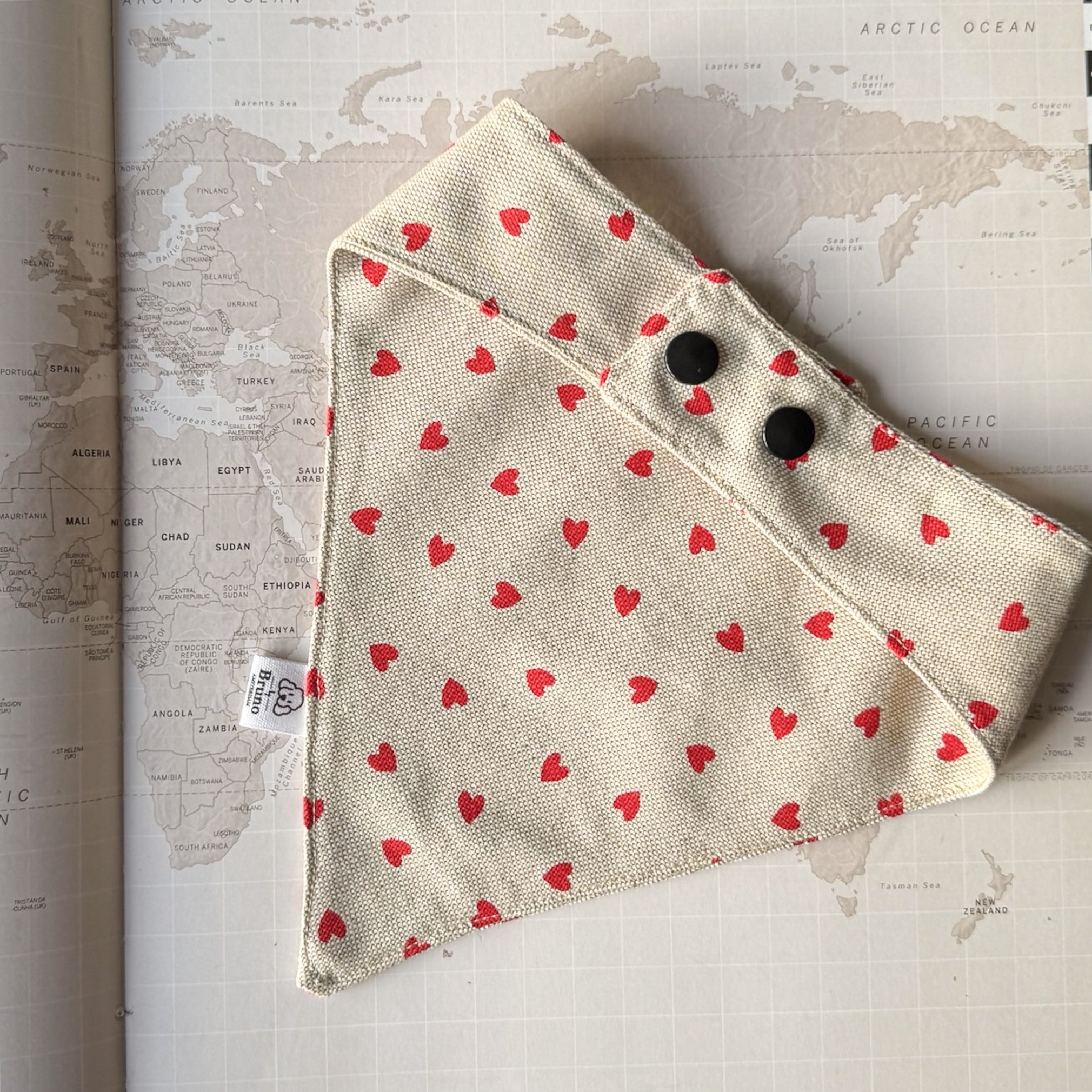 Back side of a beige dog bandana with red hearts and two black plastic buttons on a world map background.