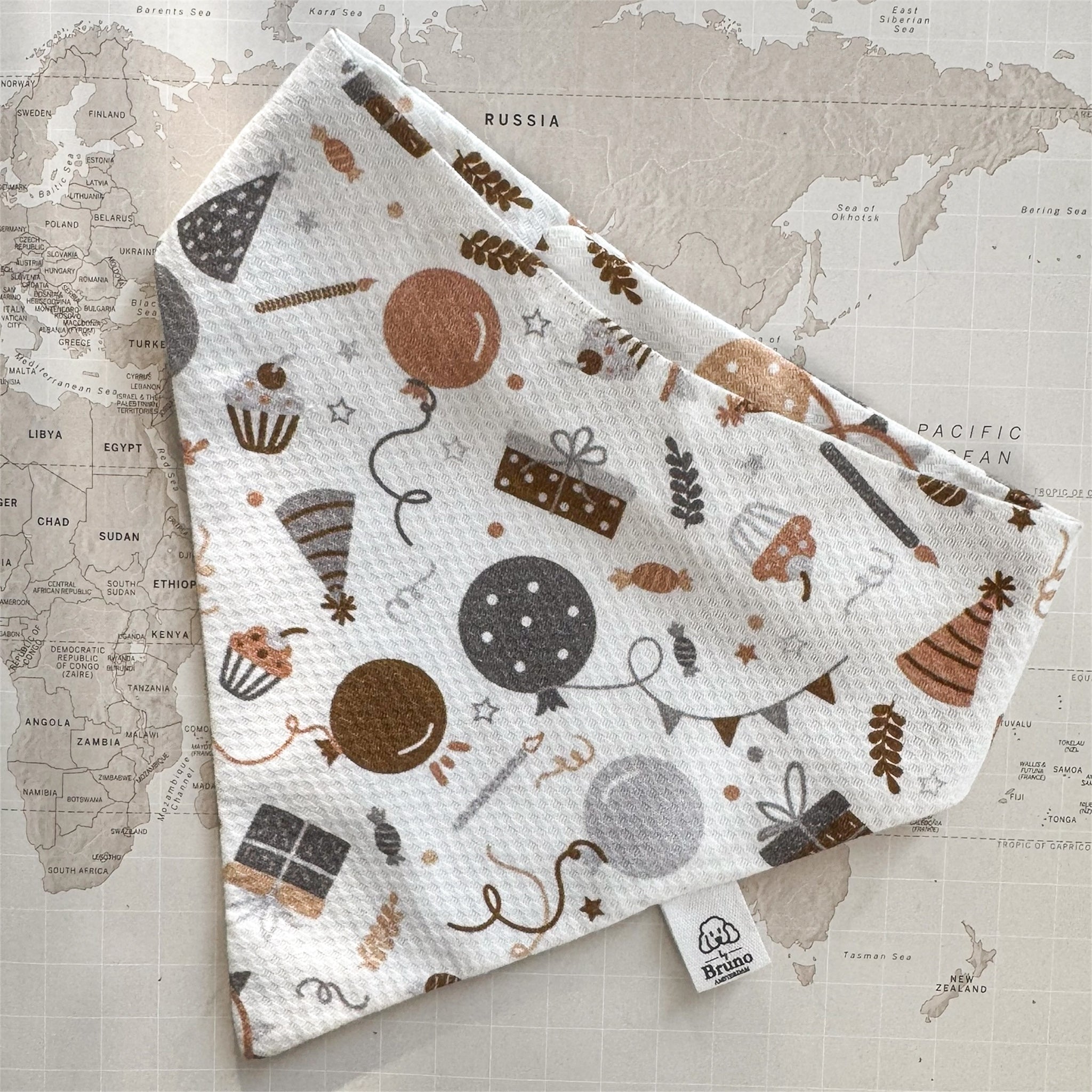 White dog bandana with balloons, wrapped gifts, cupcakes and party hats on a world map background.