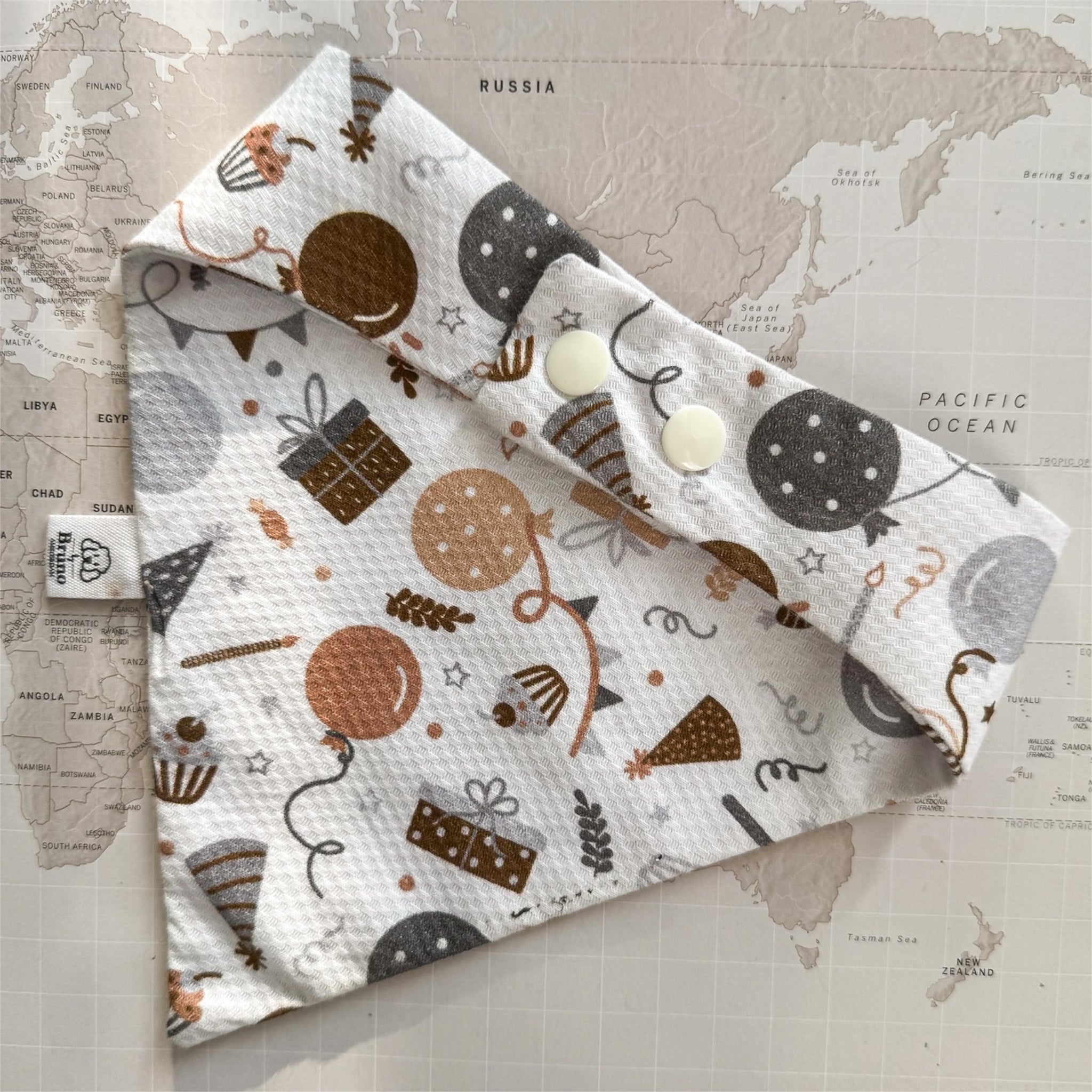 Back of a white dog bandana with balloons, wrapped gifts, cupcakes and party hats closed with peal coloured buttons and placed on a world map background.