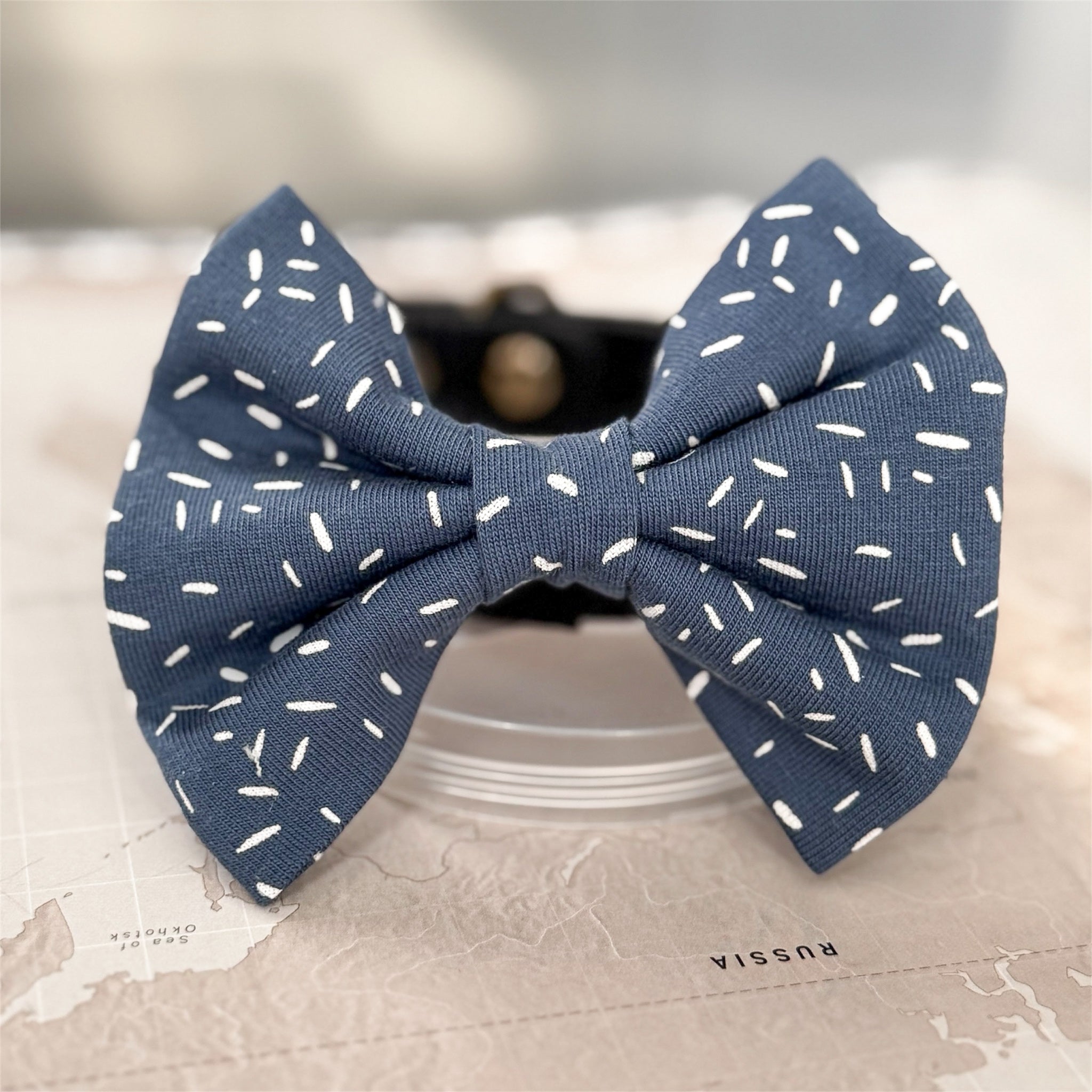 Dusty blue dog bow with white sprinkle print on a world map background.