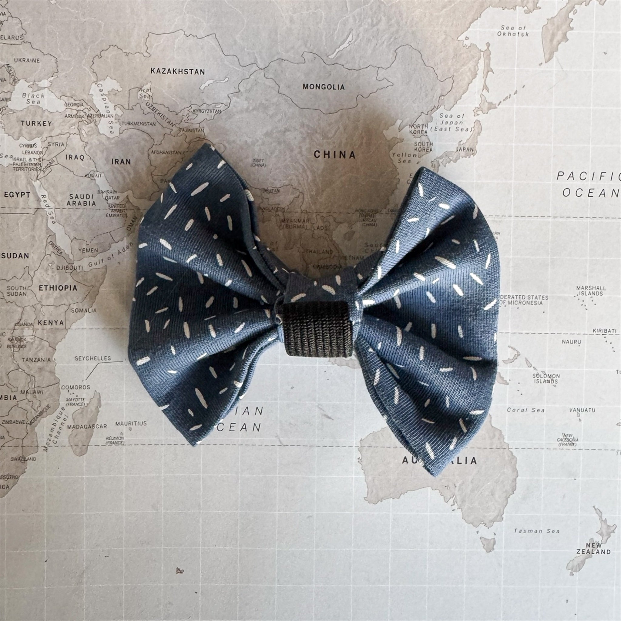 Back side of the dusty blue dog bow with white sprinkle print and an elastic loop on a world map background.