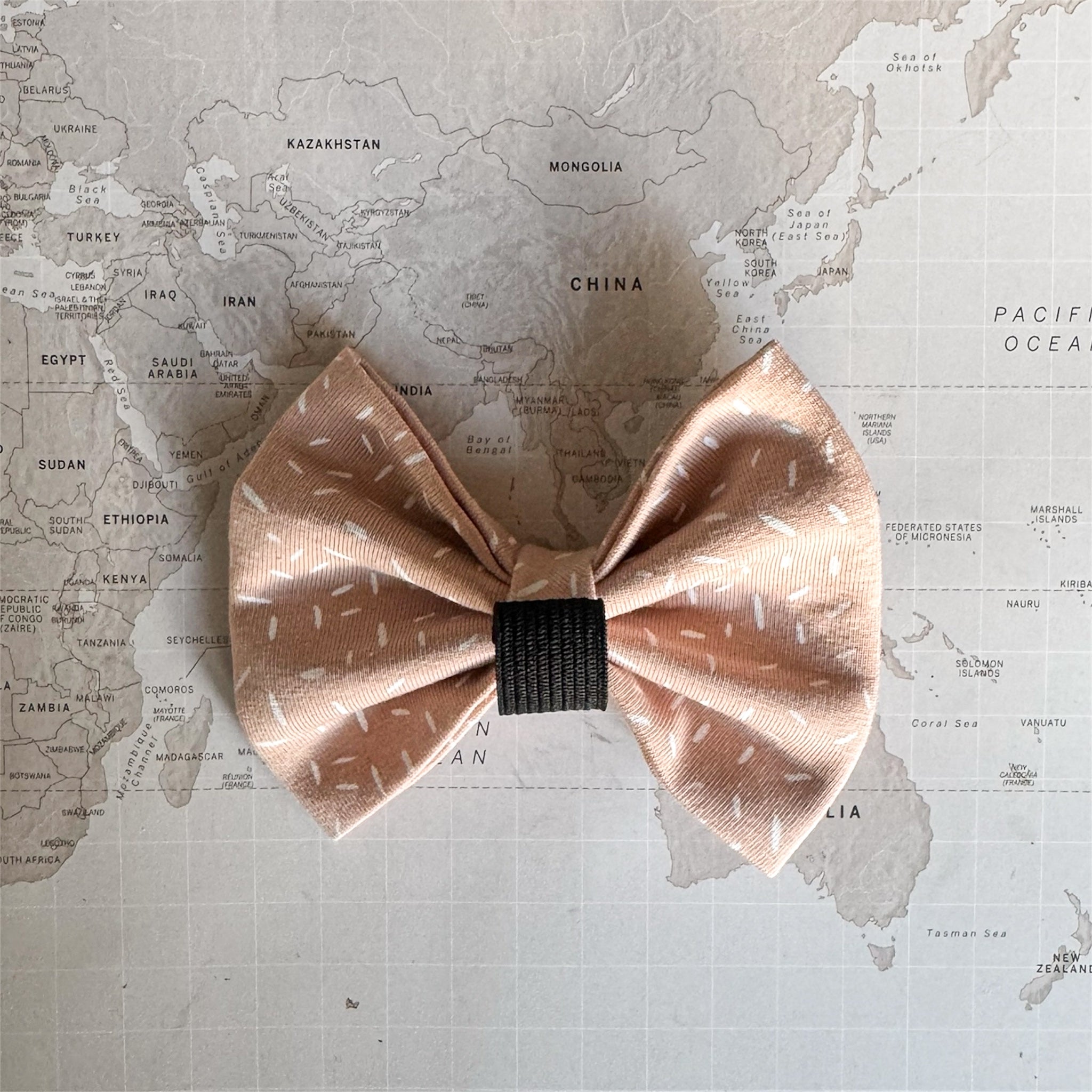 Back side of a warm apricot dog bow with white sprinkle print and an elastic loop on a world map background.