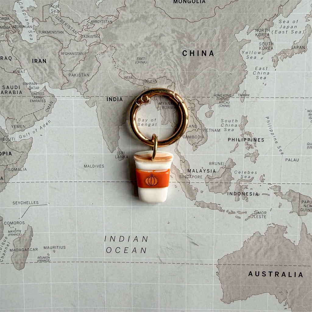 A dog tag shaped like a coffee cup with a copper-toned split ring, placed on top of a world map. The charm looks like a pumpkin spice latte to go.