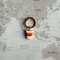 A dog tag shaped like a coffee cup with a copper-toned split ring, placed on top of a world map. The charm looks like a pumpkin spice latte to go.