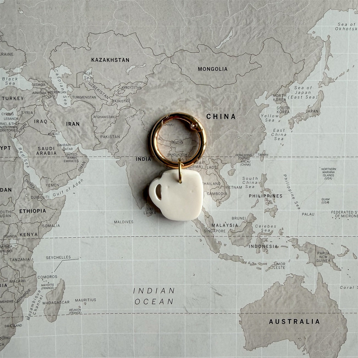 A back side of the coffee mug shaped dog tag with a gold-toned split ring, placed on top of a vintage-style world map.