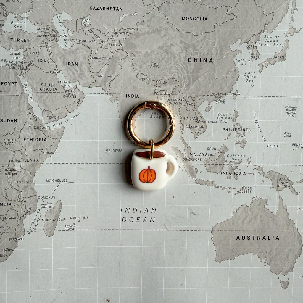 A coffee mug dog tag with an orange pumpkin design, attached to a gold split ring, displayed on a vintage-style world map background.