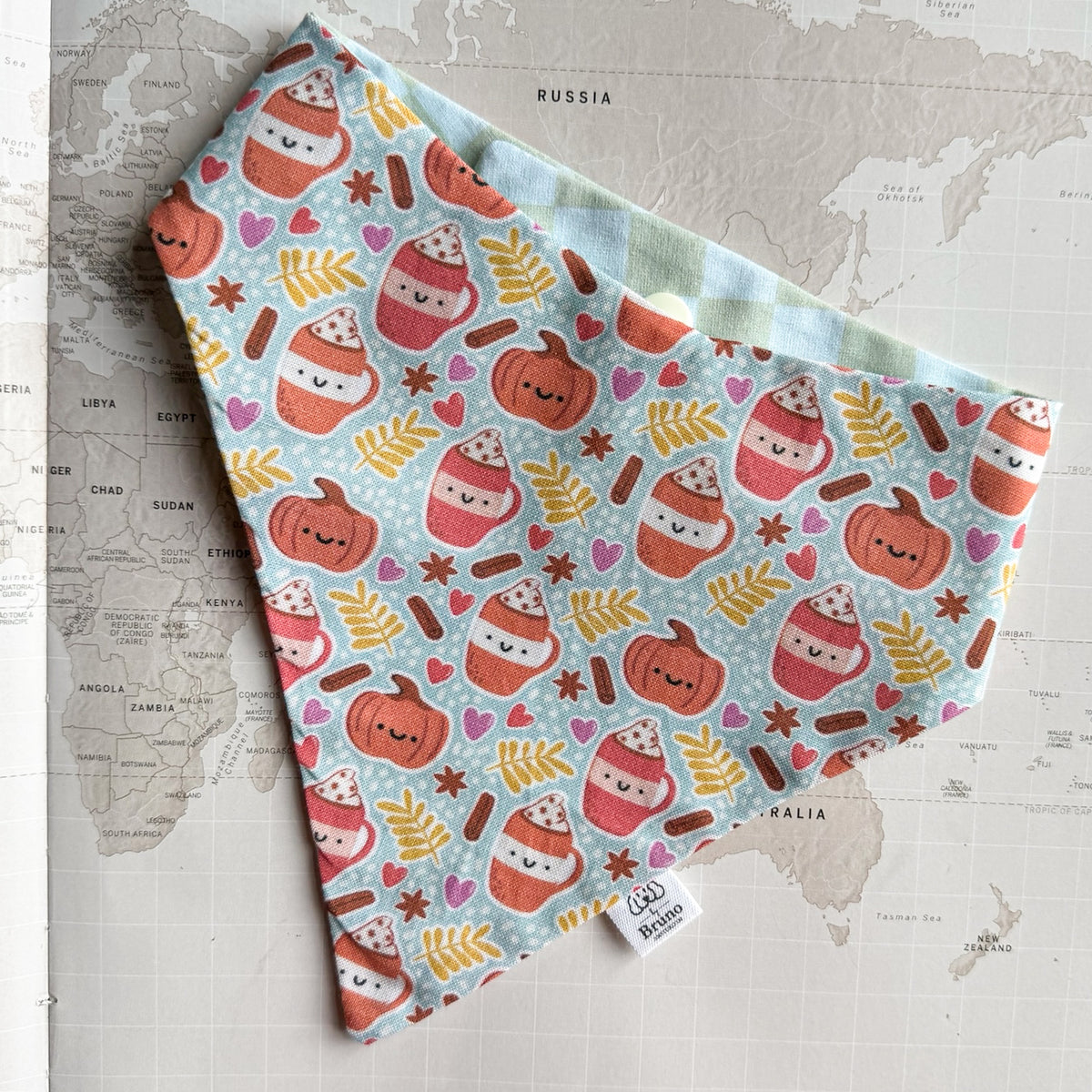 A dogbandana with a cute autumn pattern of smiling pumpkins, hot cocoa cups, leaves, and hearts on a light blue background, laid flat on a vintage world map. 