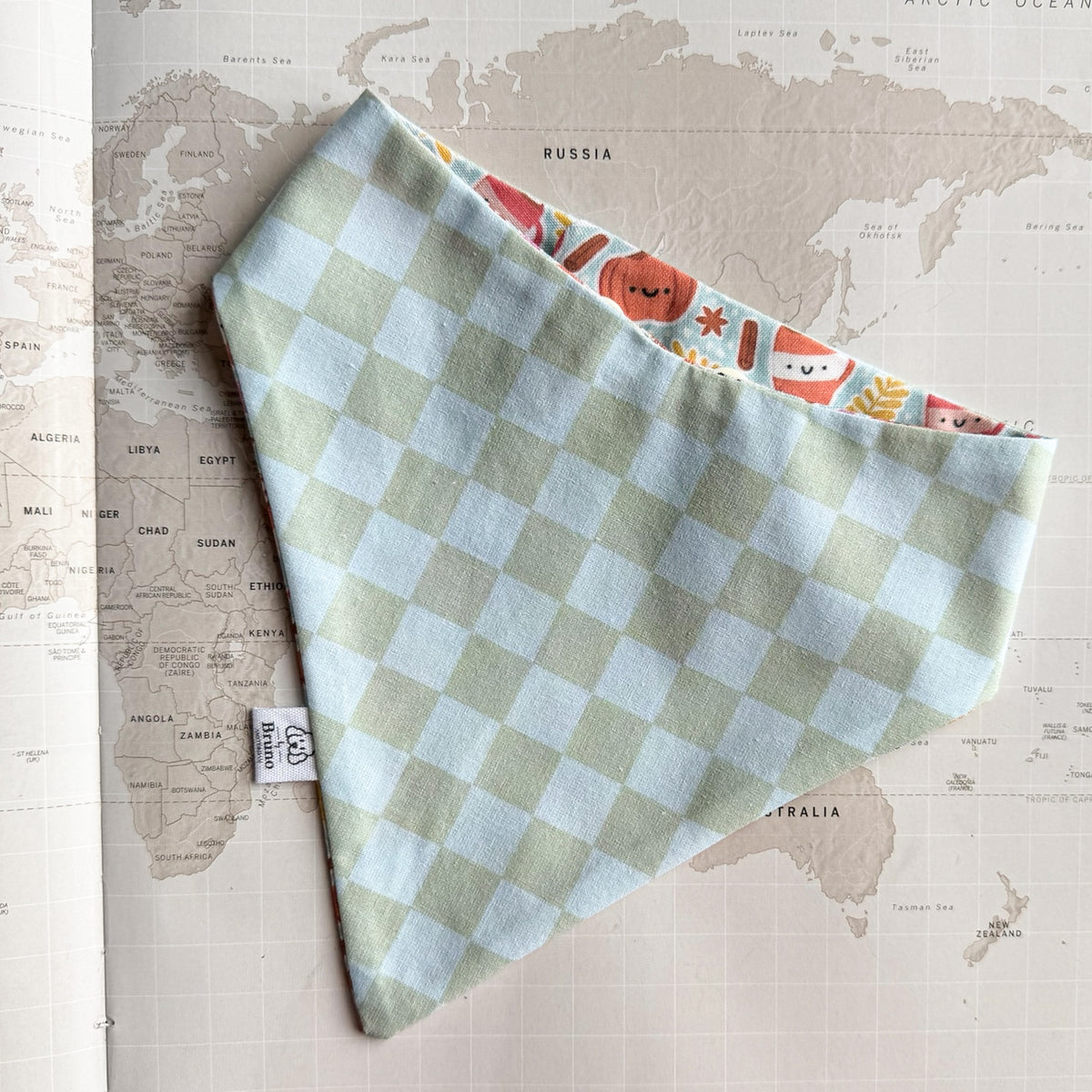 A dog bandana with a pale green and white checkered pattern, laid flat on a vintage-style world map. The inner lining has a colorful cartoon pattern with orange, red, and yellow illustrations.