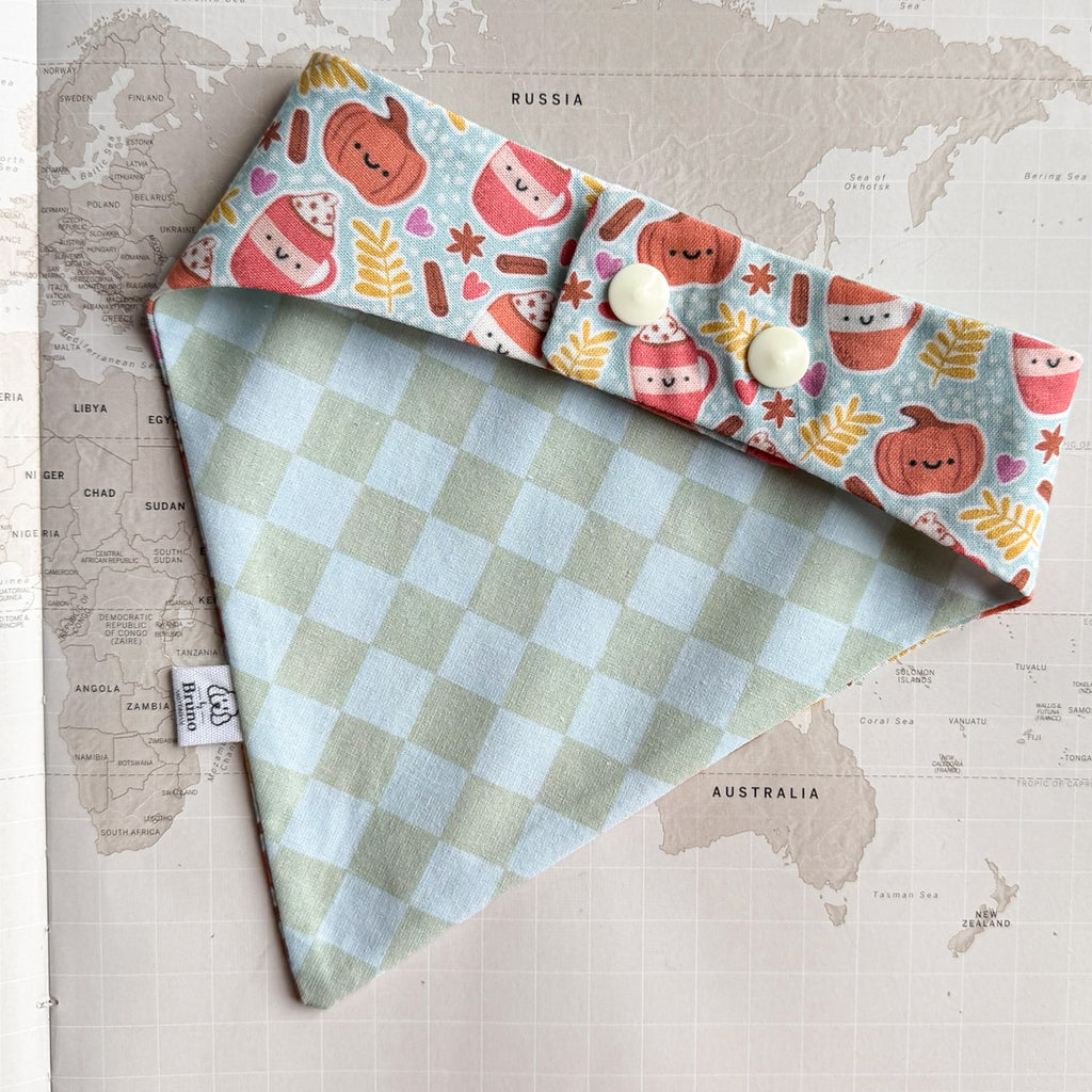 A dog bandana laid flat on a vintage world map. The fabric has a pale blue and white gingham body and a matching top band printed with cute pink and orange hot cocoa cups, pumpkins, and autumn leaves on a light blue background.