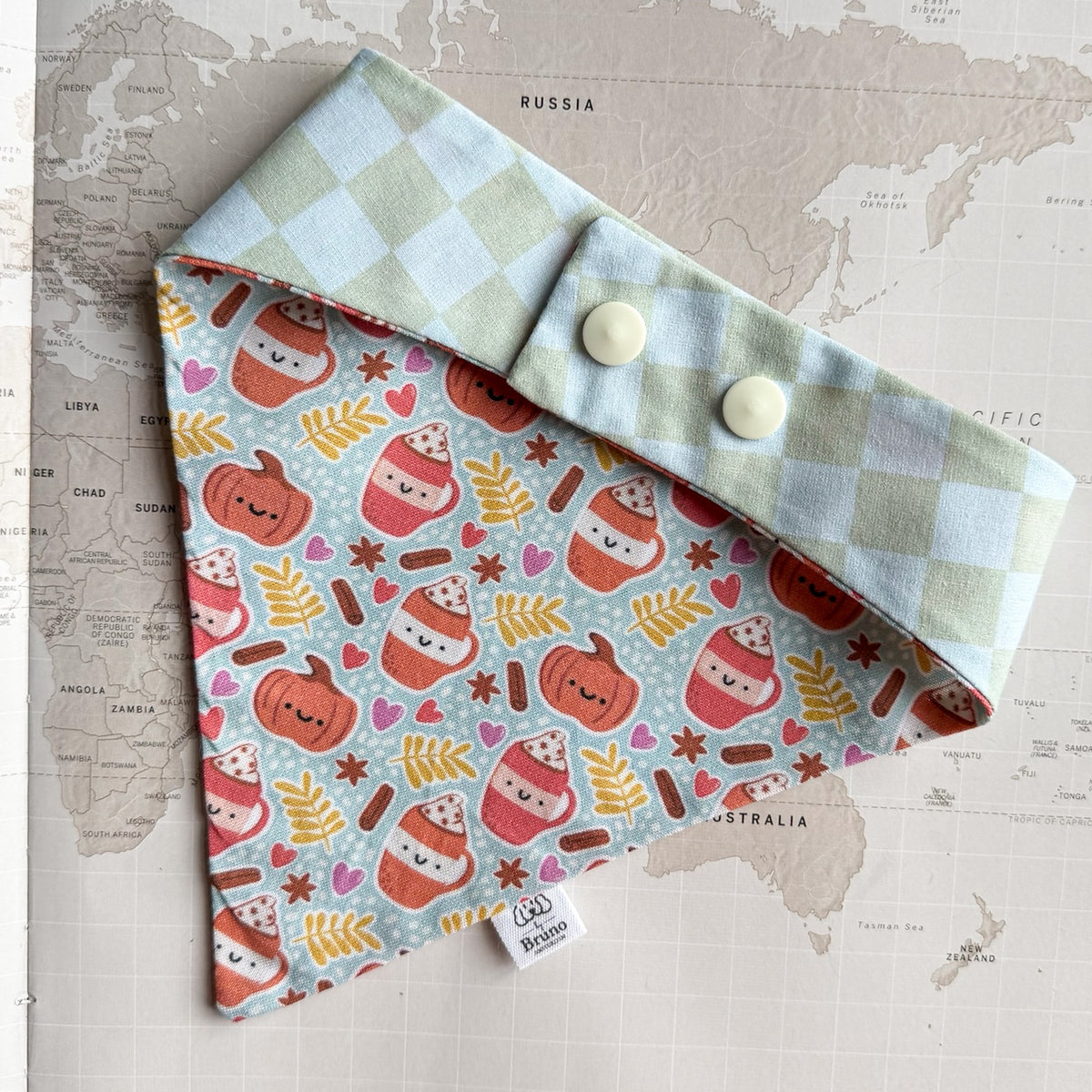 A dog bandana  laid flat on a world map background. The bandana has a pale blue and white checkered fold-over flap with two cream buttons and a front panel printed with a repeating pattern of smiling hot cocoa cups, pumpkins, leaves, and hearts on a light blue background.