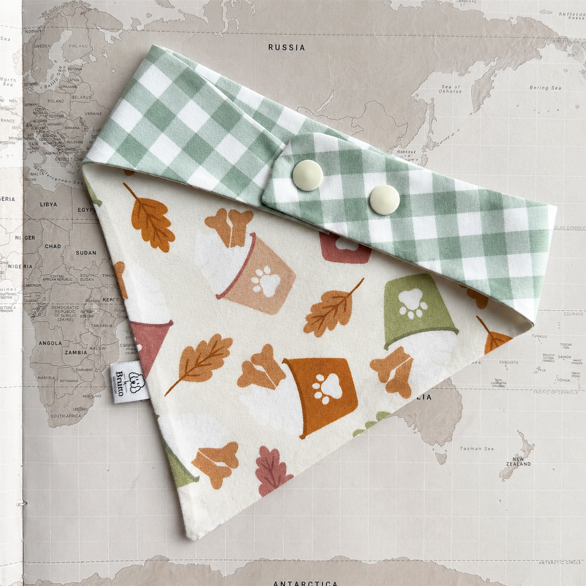 A triangular dog bandana with a green and white gingham checkered top fold and a bottom fold featuring a pattern of autumn leaves, paw prints, and dog houses on an off-white background. The bandana is styled on top of a vintage-style world map.