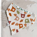 A triangular dog bandana with a white base, printed with autumn leaves, paw prints, and colorful cup motifs, laid flat on a vintage-style world map background.