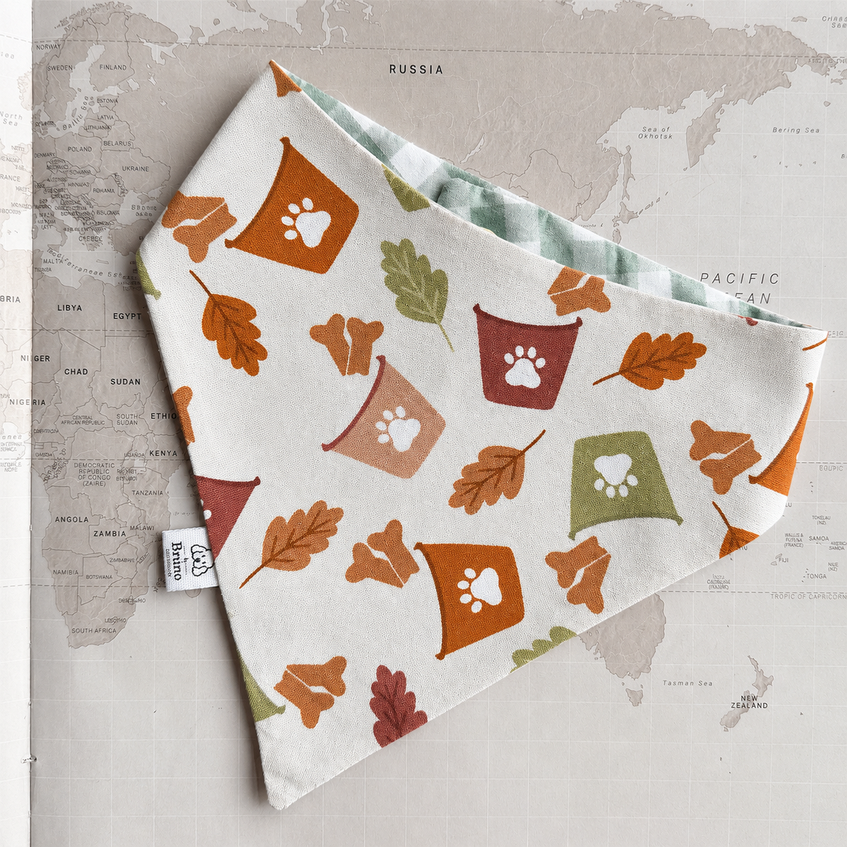 A triangular dog bandana with a white base, printed with autumn leaves, paw prints, and colorful cup motifs, laid flat on a vintage-style world map background.