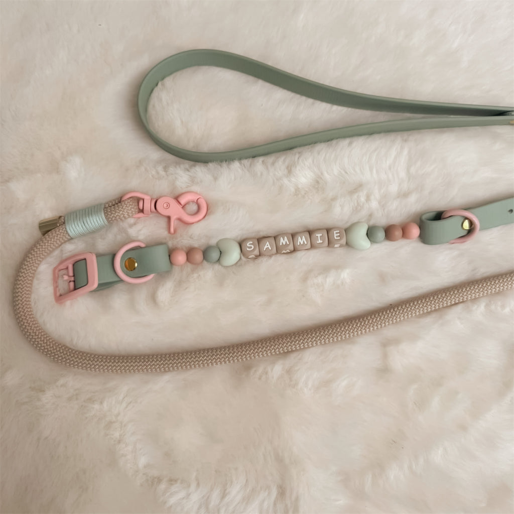 A pastel-colored pet leash with a sage green strap, pink hardware, and a beaded collar with spelling 'SAMMIE' on silicone letter blocks, laid out on a white faux fur background.