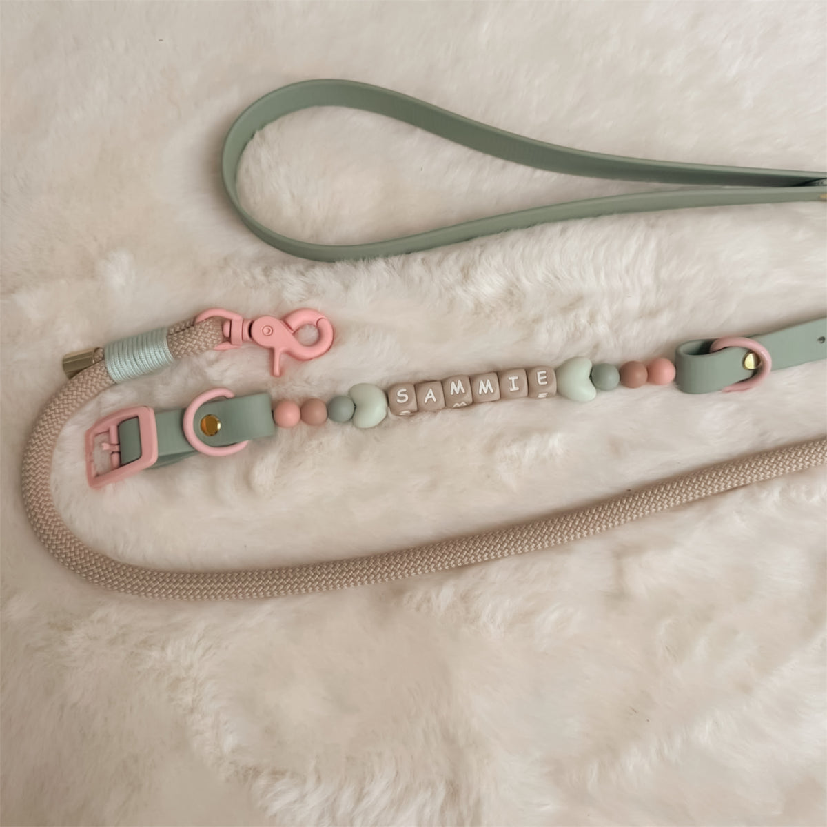 A pastel-colored pet leash with a sage green strap, pink hardware, and a beaded collar with spelling 'SAMMIE' on silicone letter blocks, laid out on a white faux fur background.