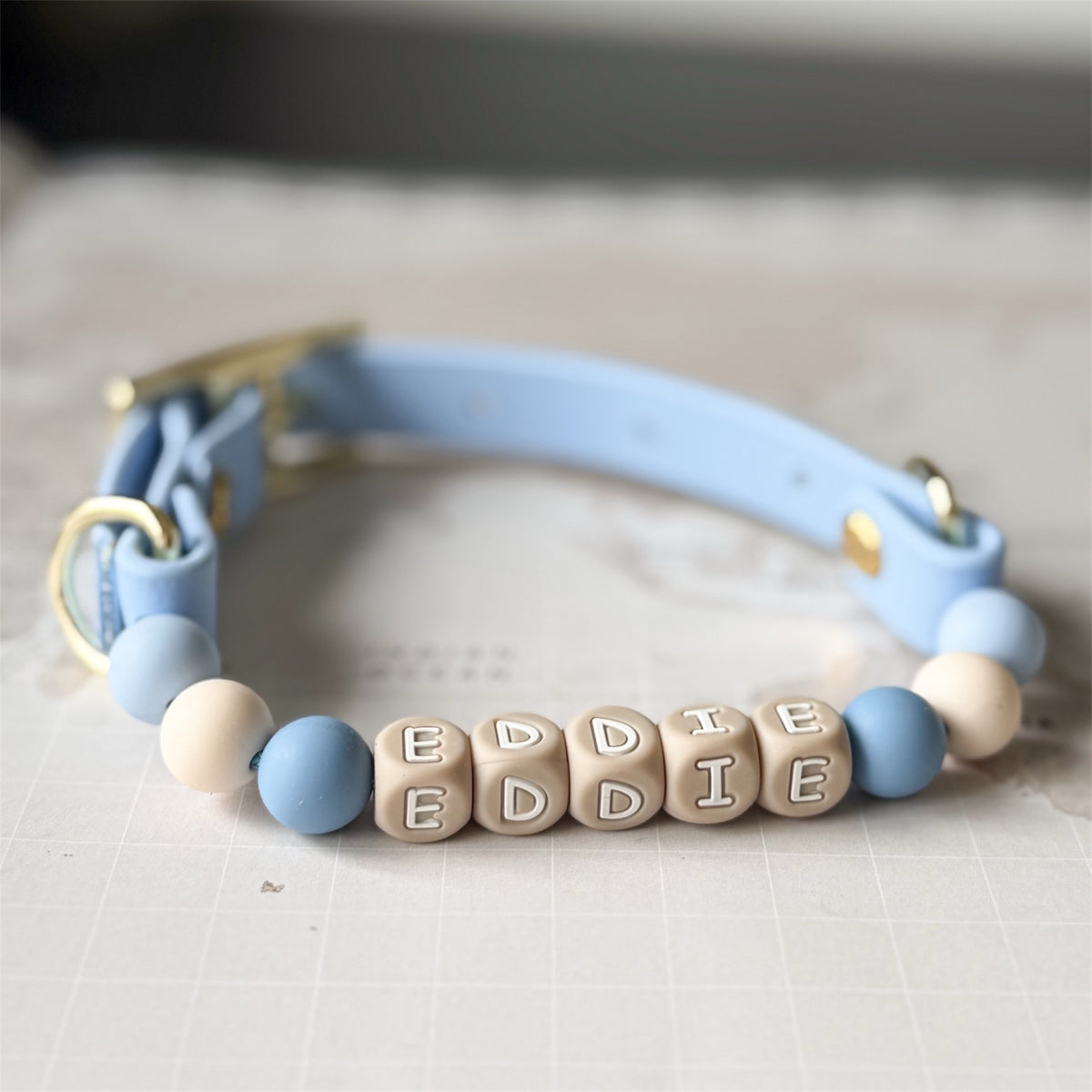 A light blue pet collar with gold-tone hardware and a beaded front section featuring wooden cube letters spelling 'EDDIE' alongside light blue and cream beads, displayed on a light surface.