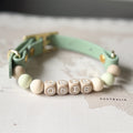 A mint green pet collar with gold-tone hardware and a beaded front. The beads include pale mint green round beads, natural wood cube beads spelling 'ODIE', and a natural wood cube bead with 'ODIE' written on it.
