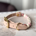 A light pink leather dog collar with gold-tone hardware and a beaded accent section featuring pink, cream, and mauve beads, resting on a vintage-style world map.