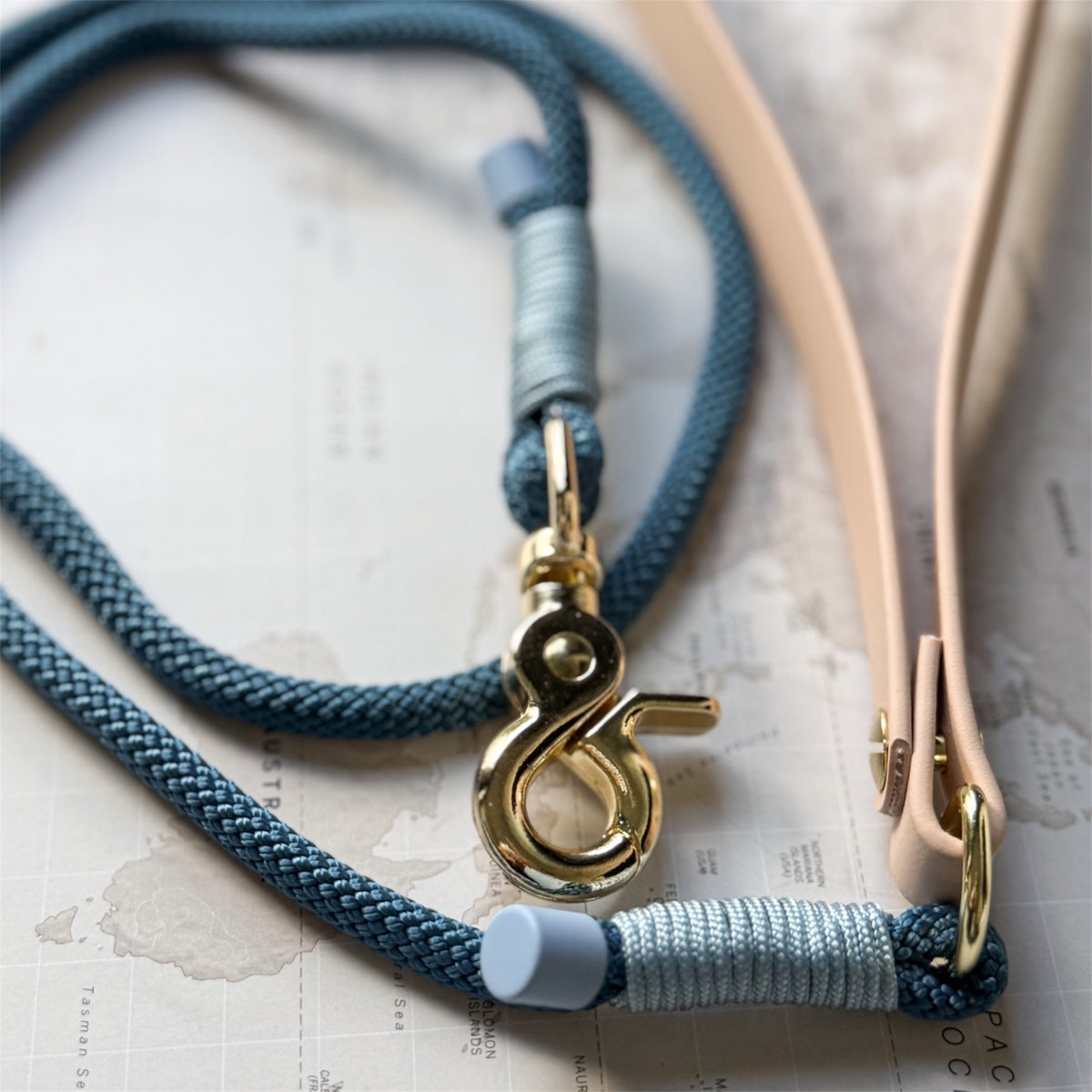 A close-up of a braided blue rope leash with a gold-tone swivel clasp and light blue wrapped cord, laid over a beige leather strap on a map background.