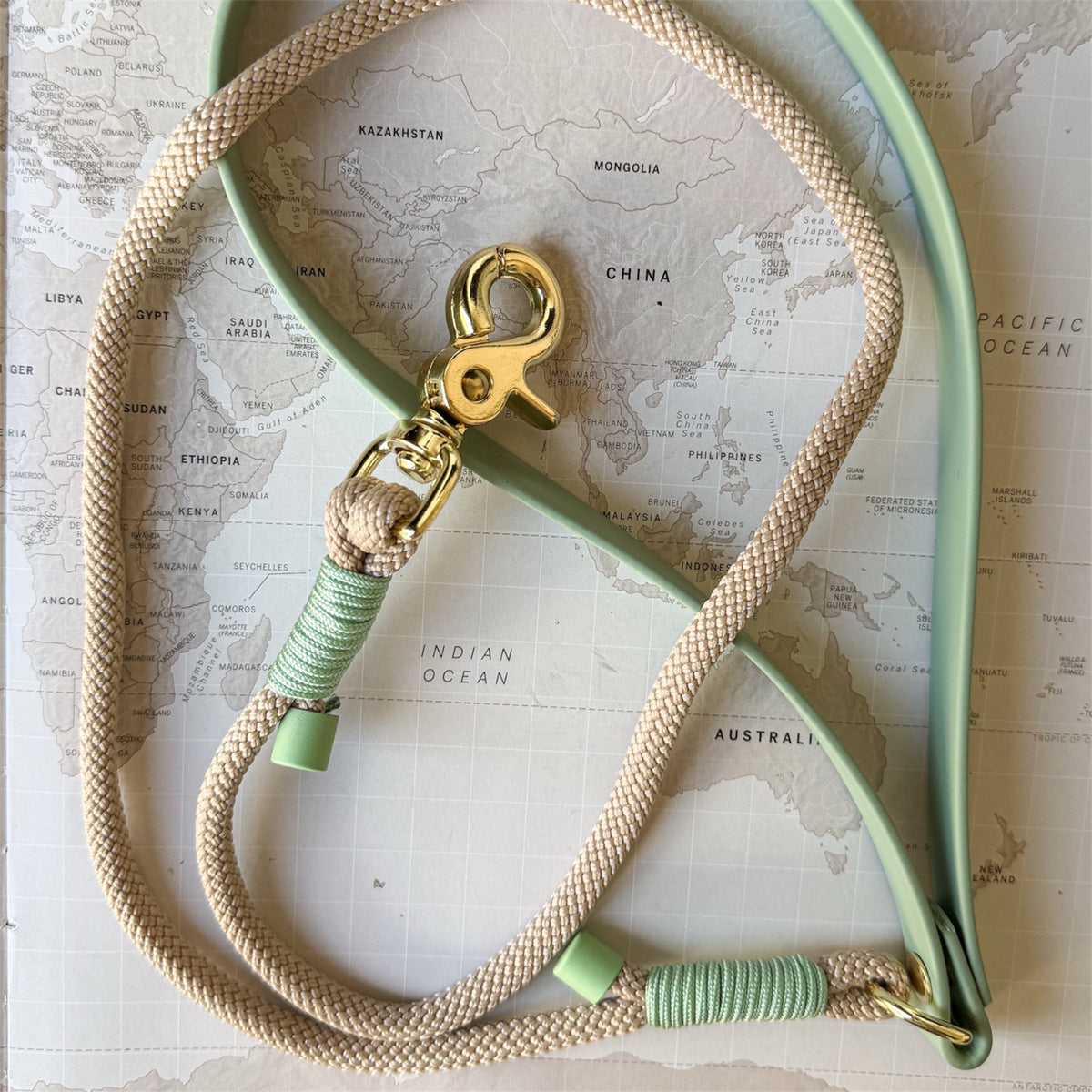 A mint green leather dog leash with a beige braided rope handle, wrapped in sage green cord, and a gold-tone lobster clasp, laid flat on a vintage-style world map.