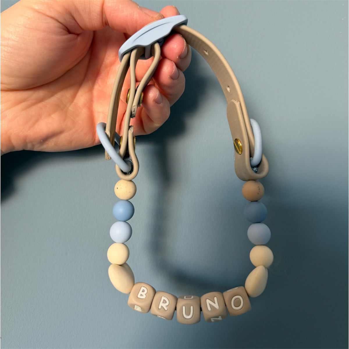 A dog collar made with biothane and silicone beads held by a hand. The collar features a beige biothane strap with light blue rings and buckle, decorated with pastel blue and beige beads, and a bottom row of beige letter cubes spelling 'BRUNO'.